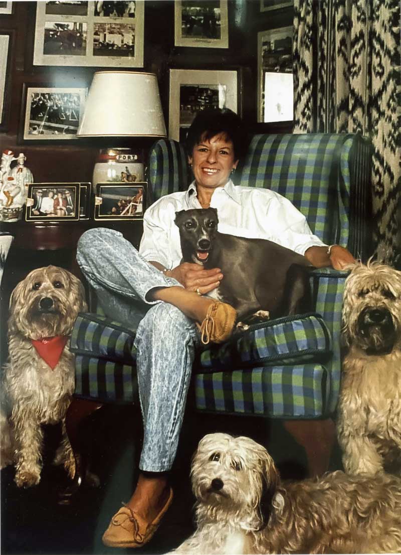Peggy Augustus seated in a plaid wingback chair surrounded by her four pet dogs.
